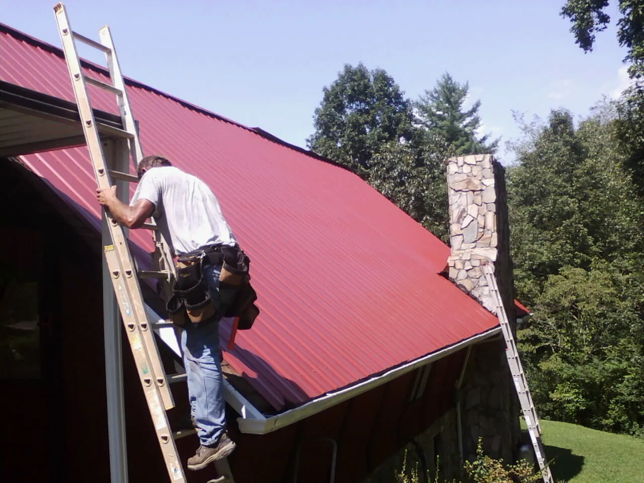 Roofer on a ladder beside a red metal roof and stone chimney — Soffit & Fascia Repair by Roofing Brothers in Wakeman